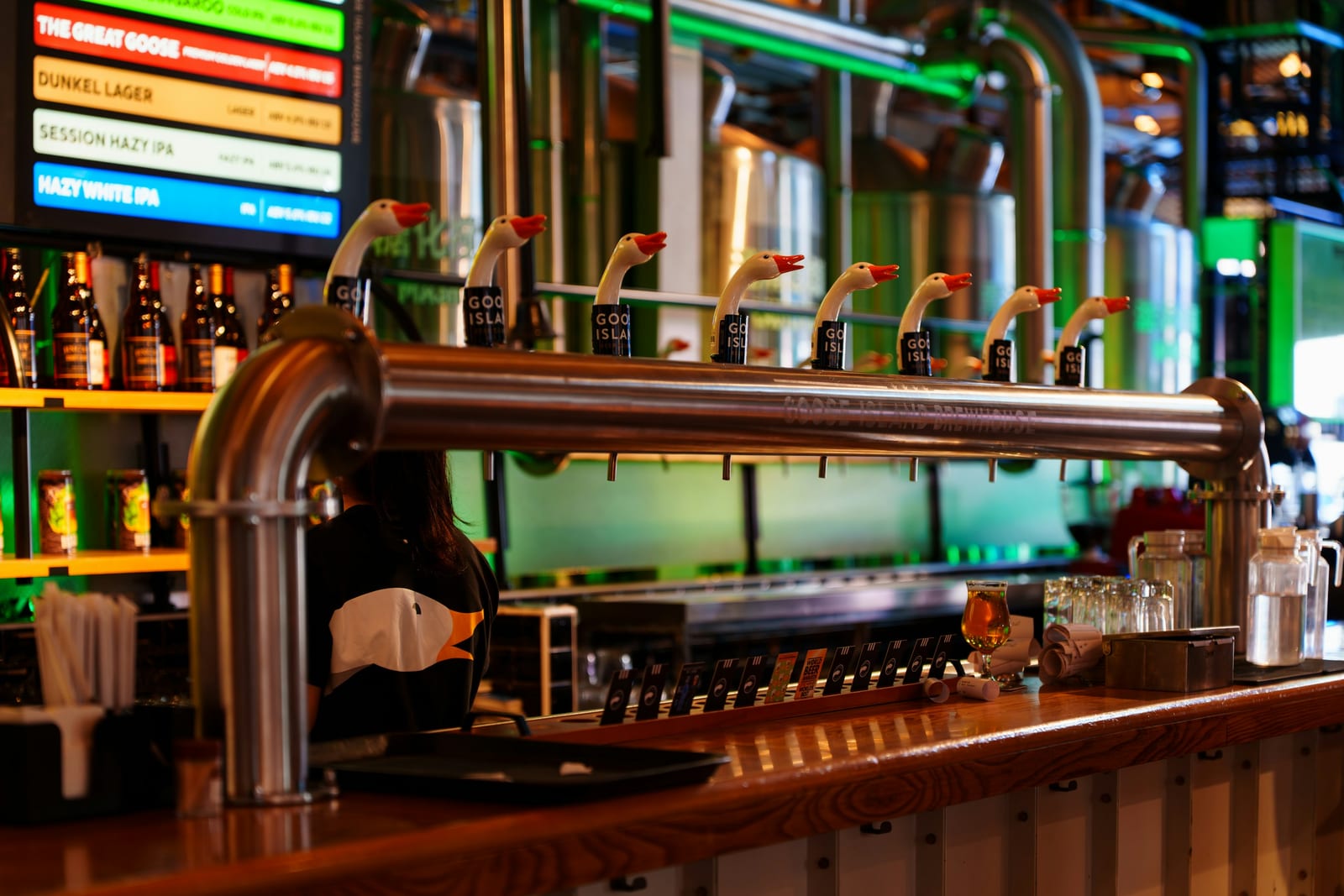 The interior of a brewery taproom with steel tanks and a long wooden bar.