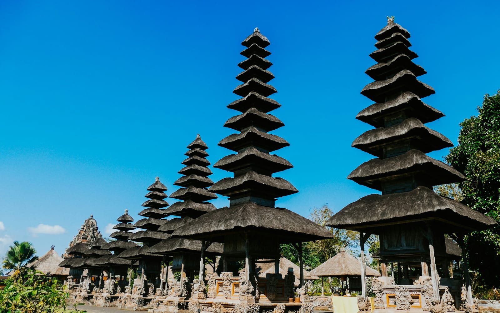 A Balinese temple garden with tiered pagodas, stone shrines, and bright blue sky.