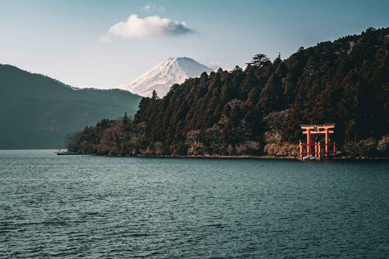 Snow-capped Mt. Fuji rises beyond Lake Ashi, with the red Hakone Shrine torii standing at the wooded shore.