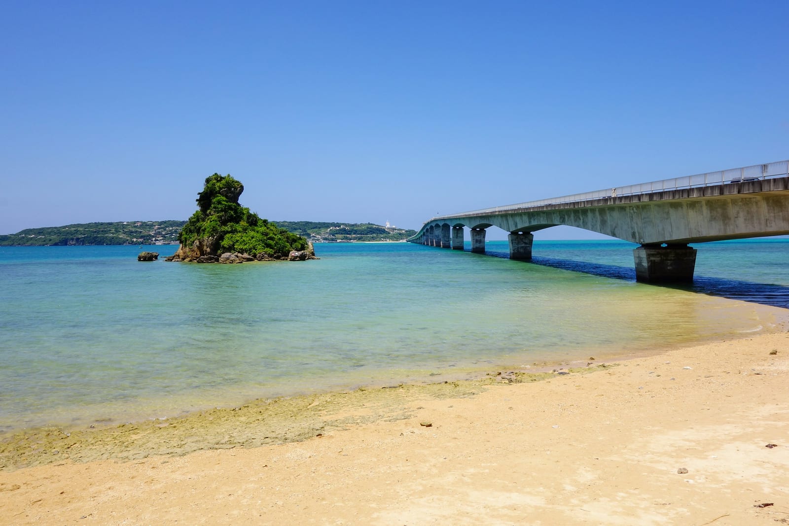 A sandy beach with clear shallow water, a small tree-covered island, and a bridge extending across the bay.