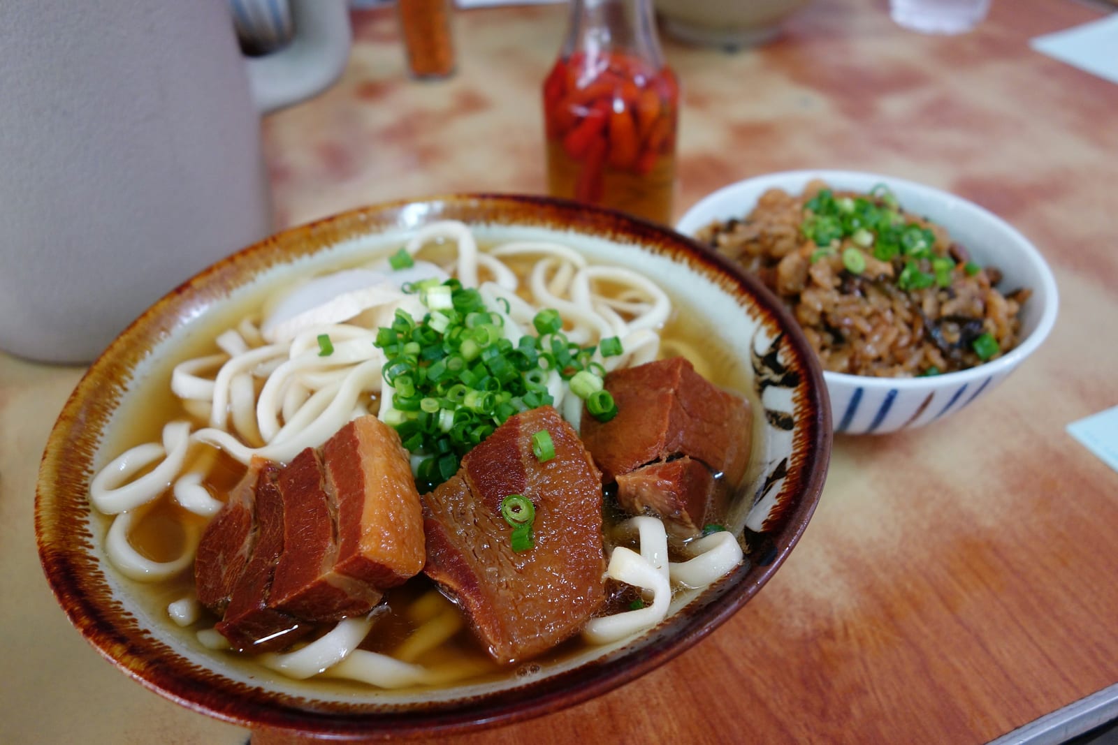 A bowl of Okinawa soba with pork, green onions, and a small side dish on a market table.