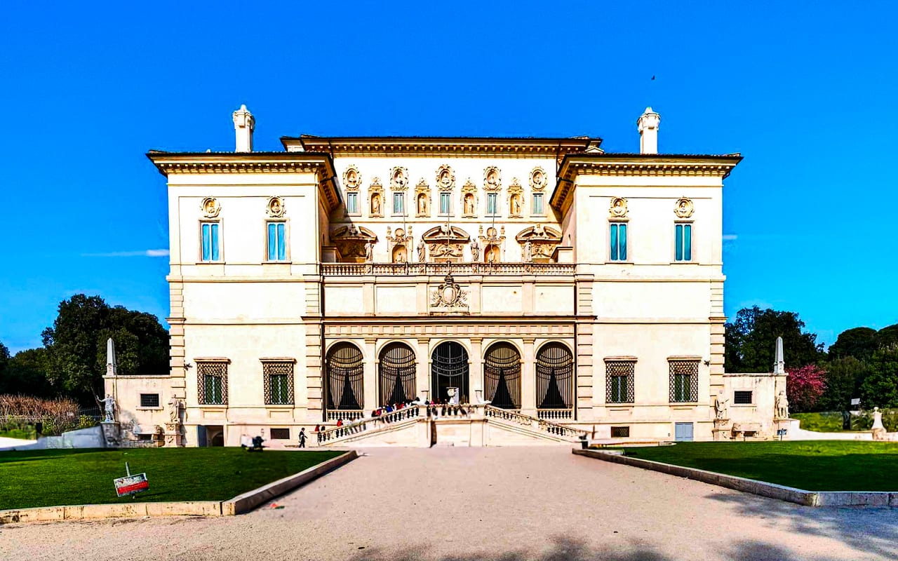 The ornate white facade of the Galleria Borghese rises above its garden avenue under a deep blue sky.