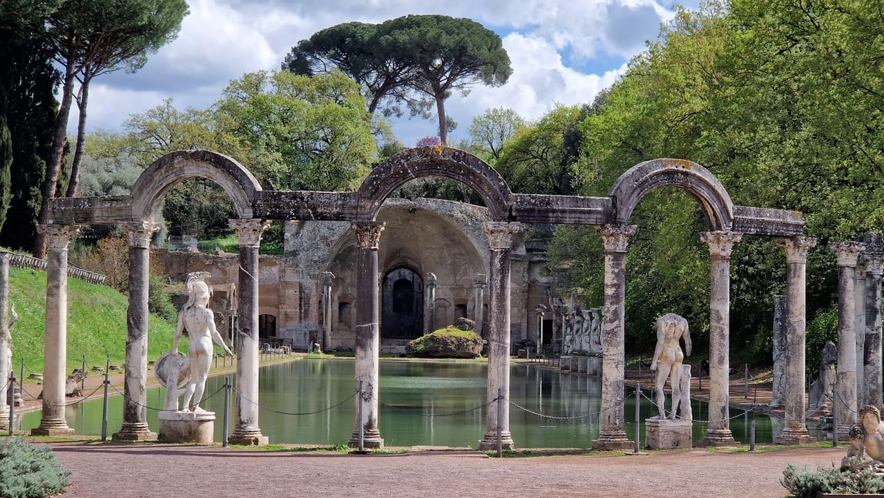 The long Canopus pool at Hadrian's Villa mirrors a curved colonnade of arches, columns, and classical statues below a Roman pine.