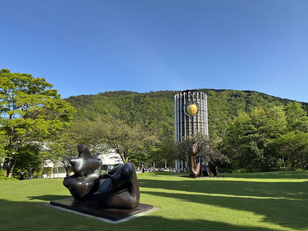 Bronze figures stand on a sunlit lawn before the museum's stained-glass Symphonic Sculpture tower, forested mountains behind.