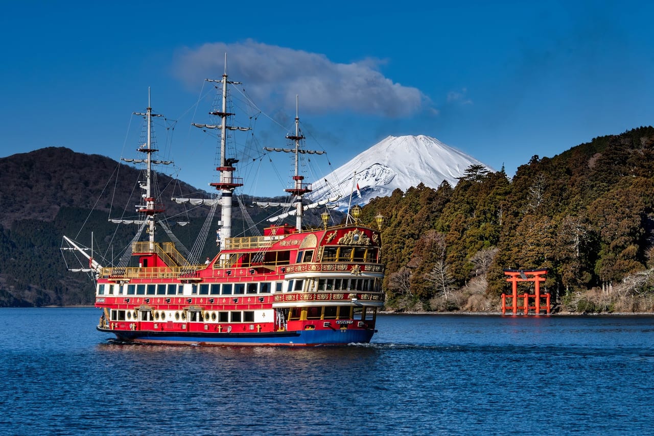 A red sightseeing galleon crosses Lake Ashi with snow-capped Mt. Fuji and a vermilion lake torii behind it.