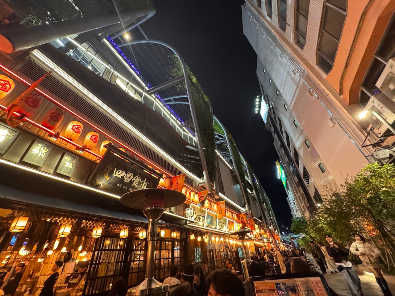 The tiny lantern-lit bars of Nonbei Yokocho line a narrow lane at night, with a modern Shibuya tower rising behind.