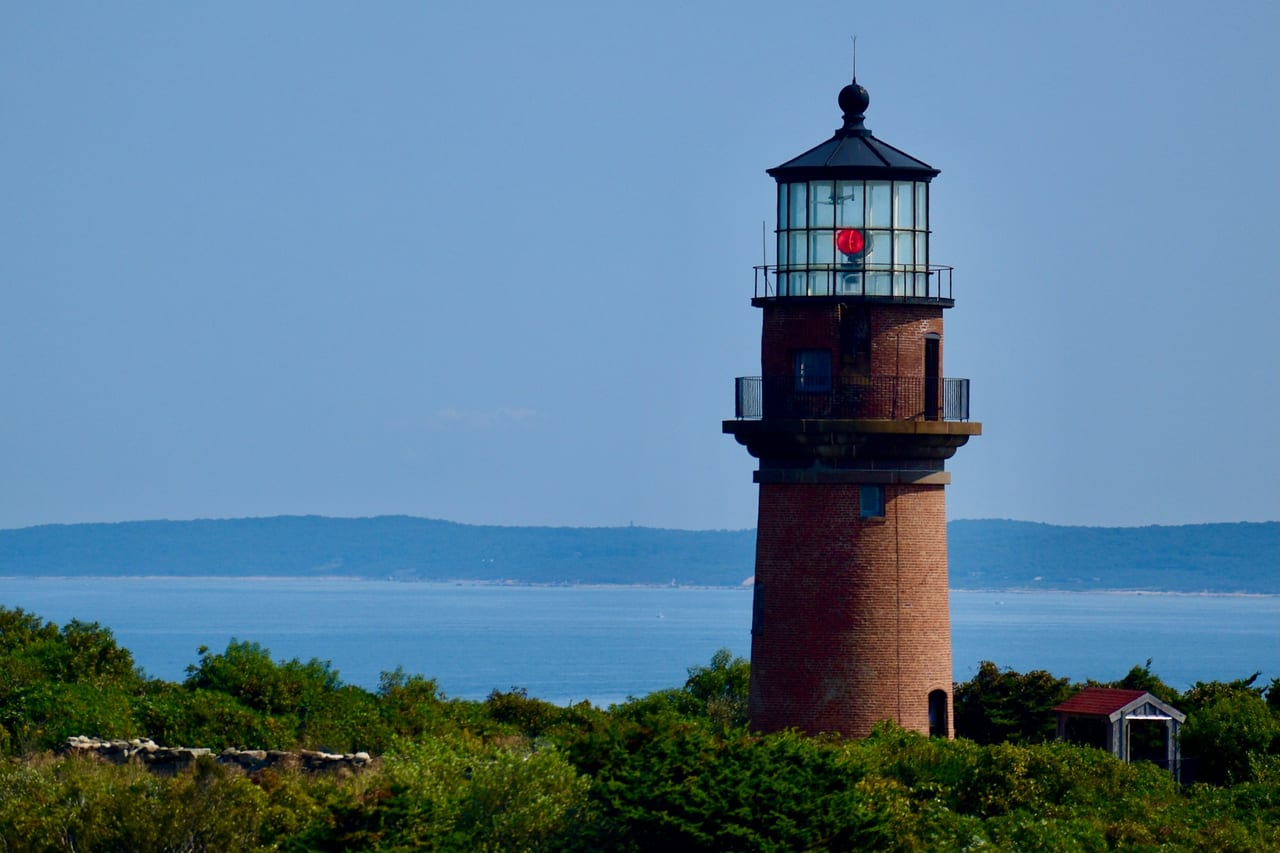 A brick lighthouse stands above trees with blue water and a distant shoreline behind it.