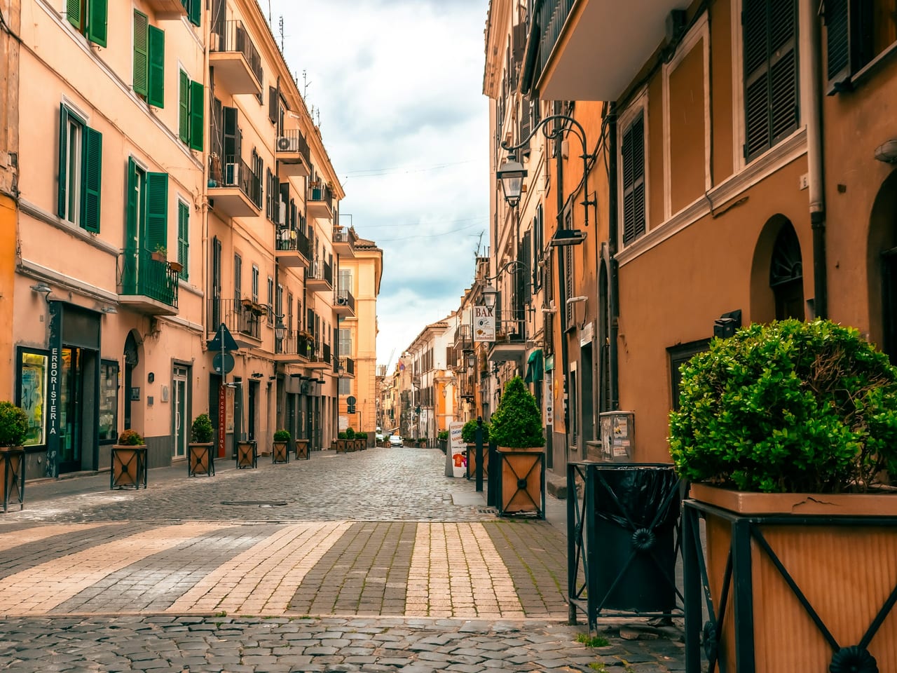 A sunlit, empty cobbled lane runs between warm ochre houses with green shutters in Rome's old center.