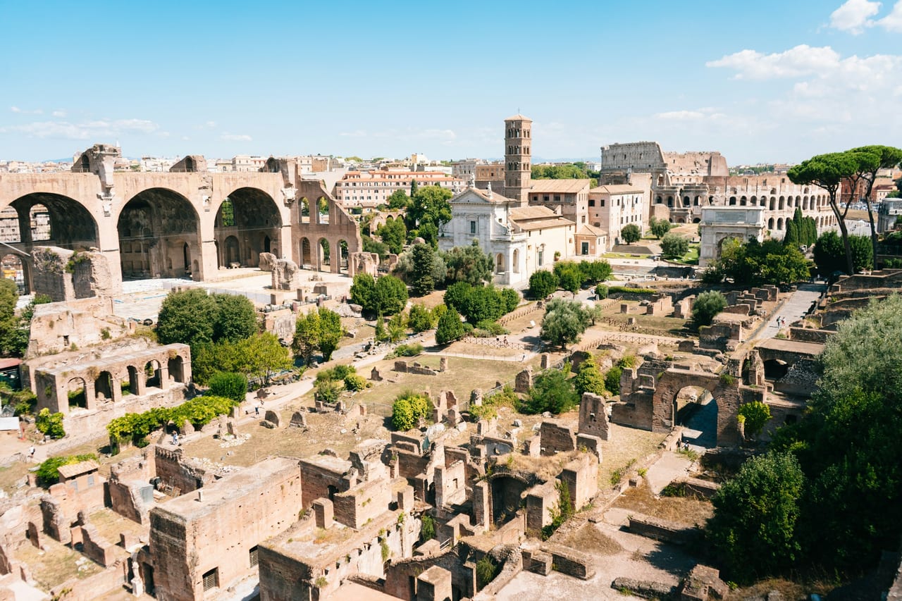 A wide view over the ruins of the Roman Forum, columns and brick arches running toward the Colosseum in the distance.