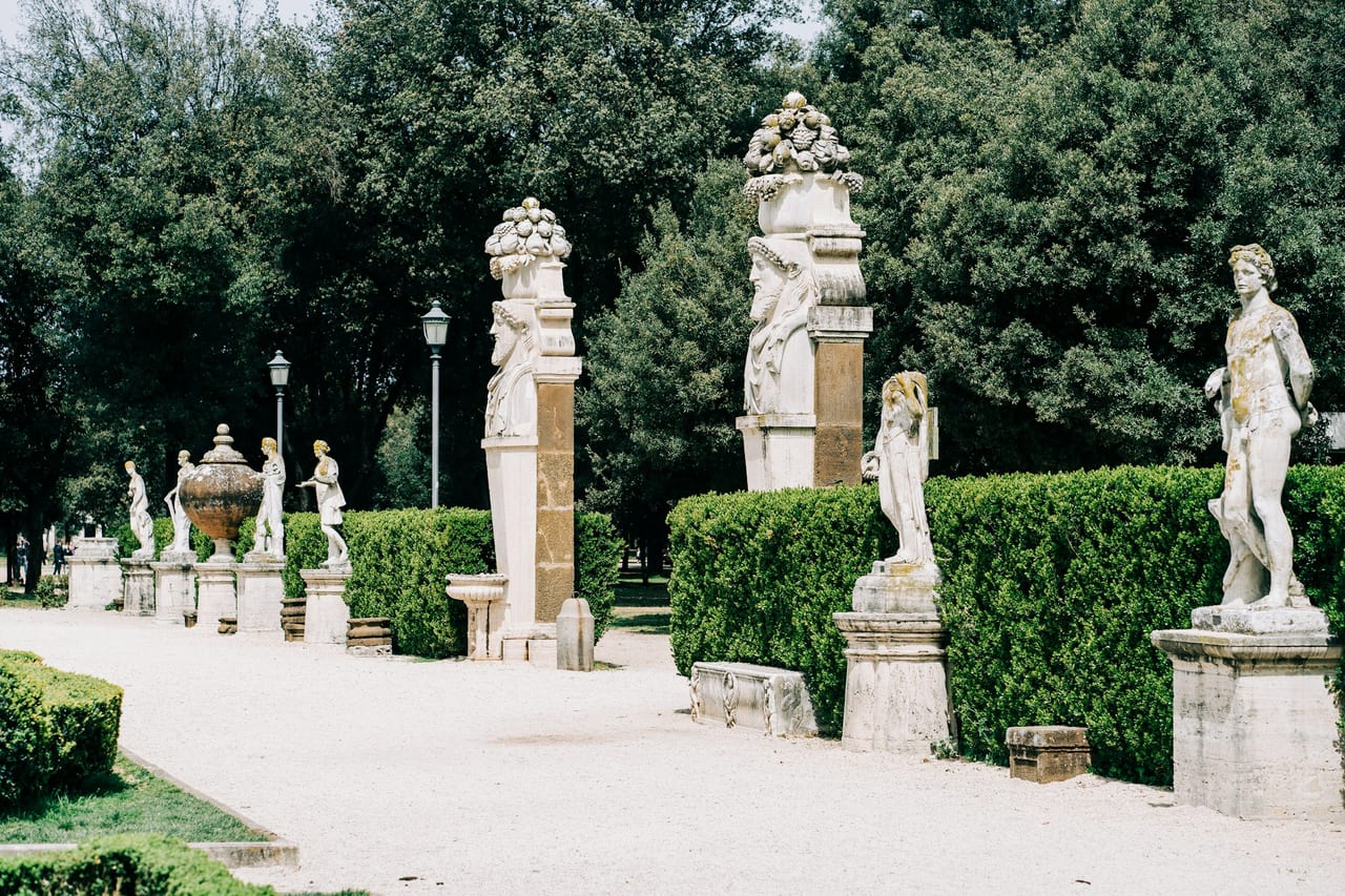 Classical marble statues line a gravel avenue among the trees of Rome's Villa Borghese gardens.