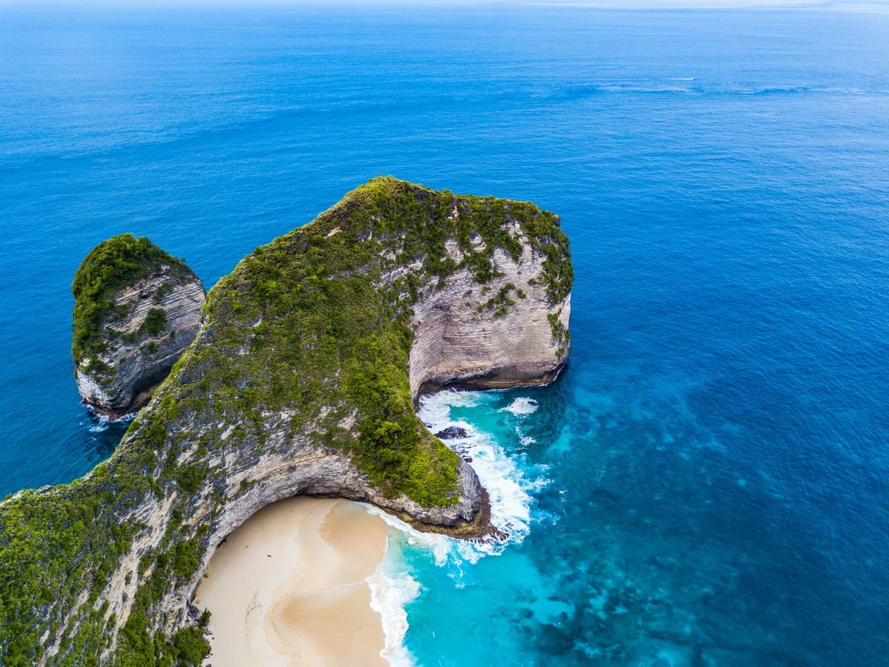 Aerial view of a rocky green headland and crescent beach surrounded by blue sea in Bali.