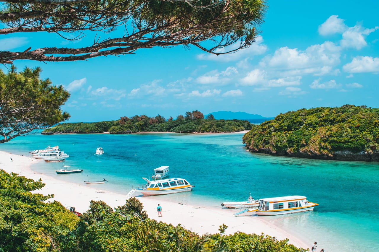 A sandy cove with small boats, turquoise water, and tree-covered islands.