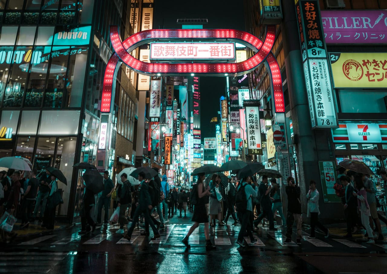 The red neon arch of Shinjuku's Kabukicho gate glows over a crowded, rain-slicked street at night.