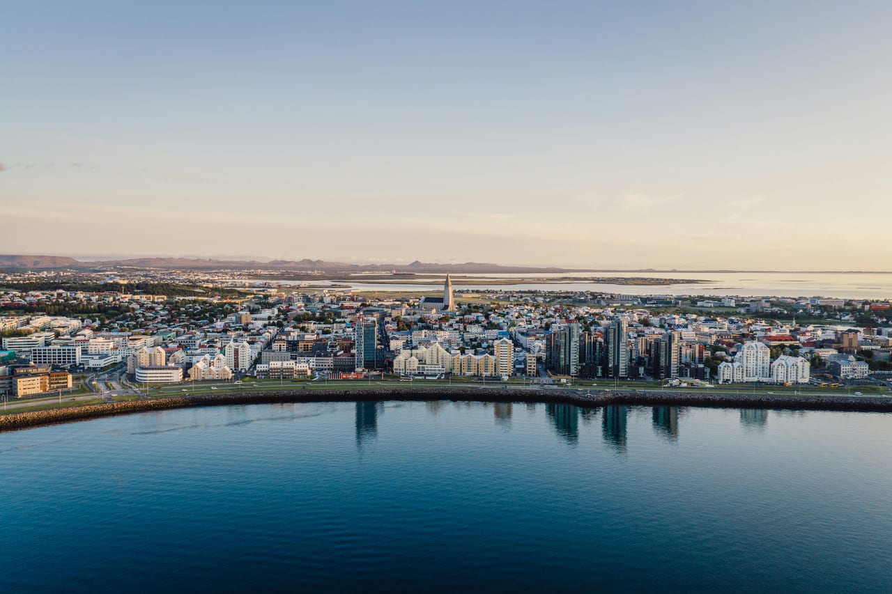 A broad view of Reykjavik across a calm waterfront with the city and distant landscape behind it.